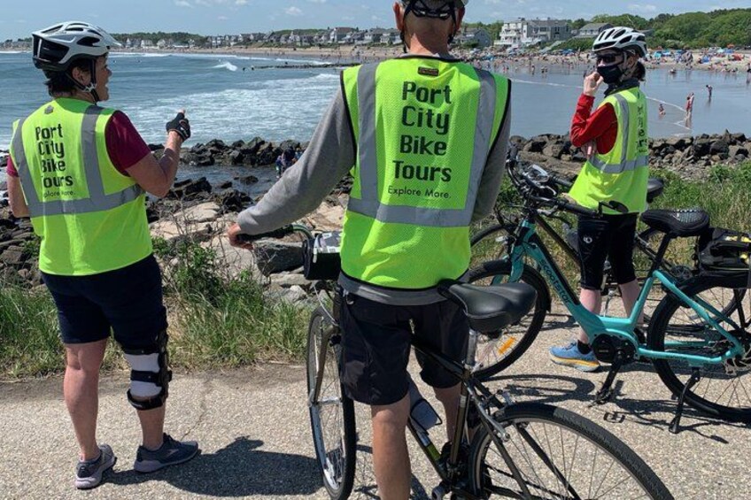 Tre personer som bär varselvästar, en av dem sitter på en cykel, och en stenstrand syns i bakgrunden. 5