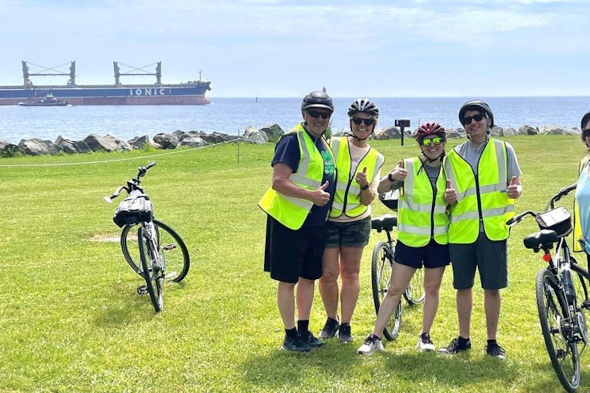 A group of people wearing safety vests standing on grass near a body of water with a cargo ship in the background. 5