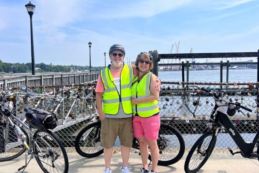 Two people standing in front of a bridge with a chain-link fence decorated with padlocks. 2
