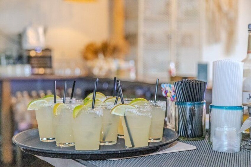 A tray of drinks with straws and lime wedges, a stack of plastic cups, and a bar setting in the background. 3
