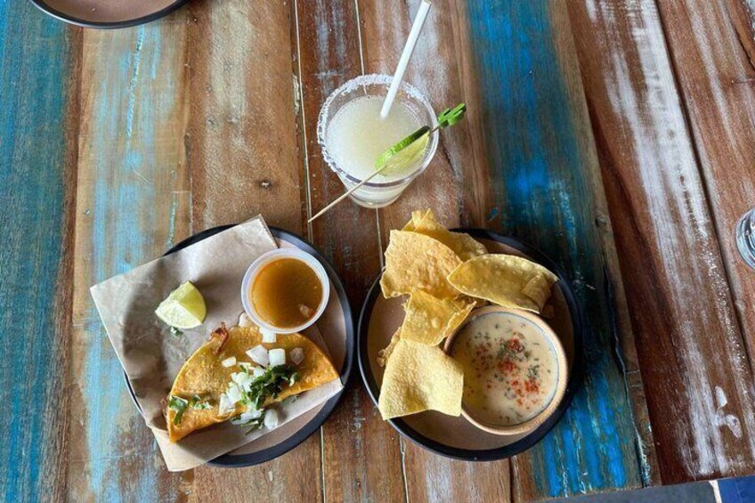 A wooden table with a plate of tacos, a bowl of salsa, a glass of a beverage with a lime wedge, and a tortilla chip bowl. 5
