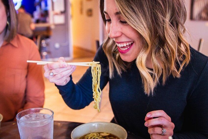 A person holding noodles with chopsticks, a glass of water, and a bowl of noodles on a table. 2