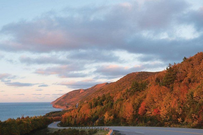 A winding road through a hilly landscape with autumn foliage, a body of water, and a cloudy sky. 1