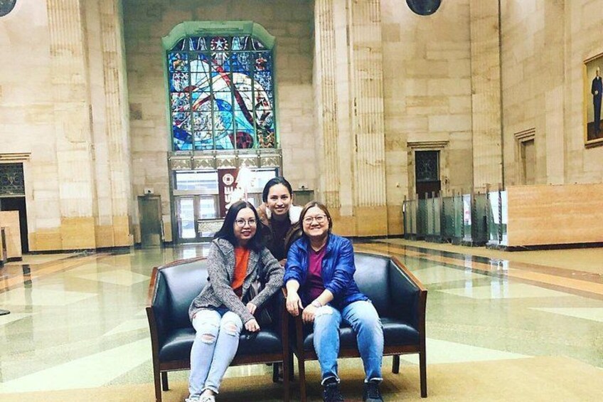Tres personas sentadas en un banco en un amplio salón con un gran ventanal de vitral al fondo. 3