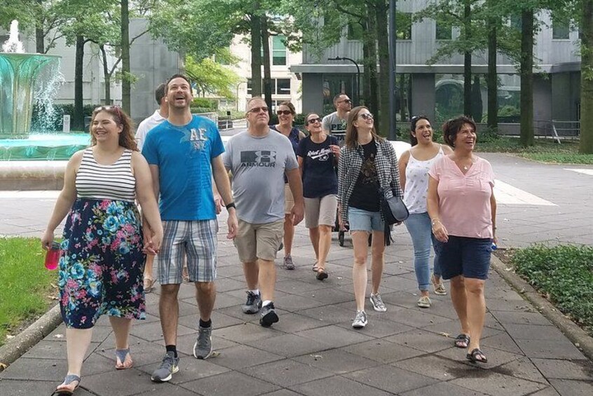 A group of people walking on a paved path in a park with trees and a fountain in the background. 1