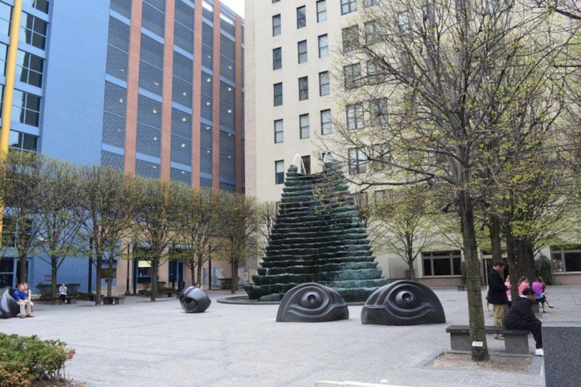 A plaza with a modern sculpture, trees, benches, and buildings in the background. 4