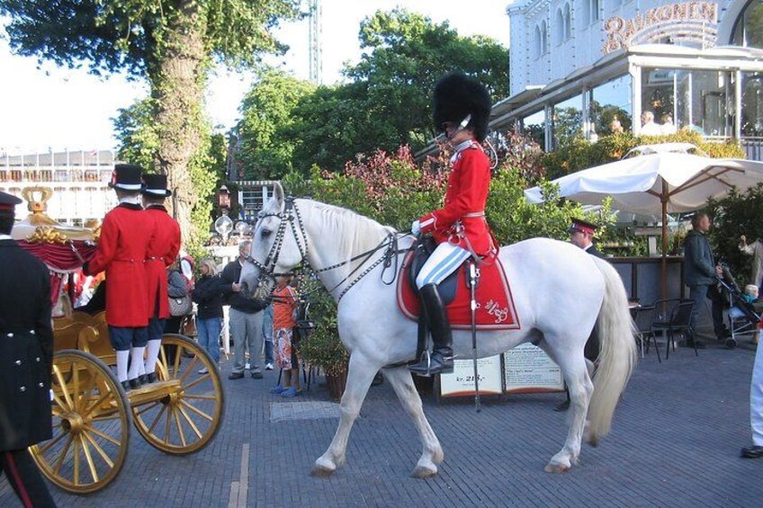Een koets getrokken door een wit paard, met een ruiter in rode uniform, en een gebouw met een borduur in de achtergrond. 5