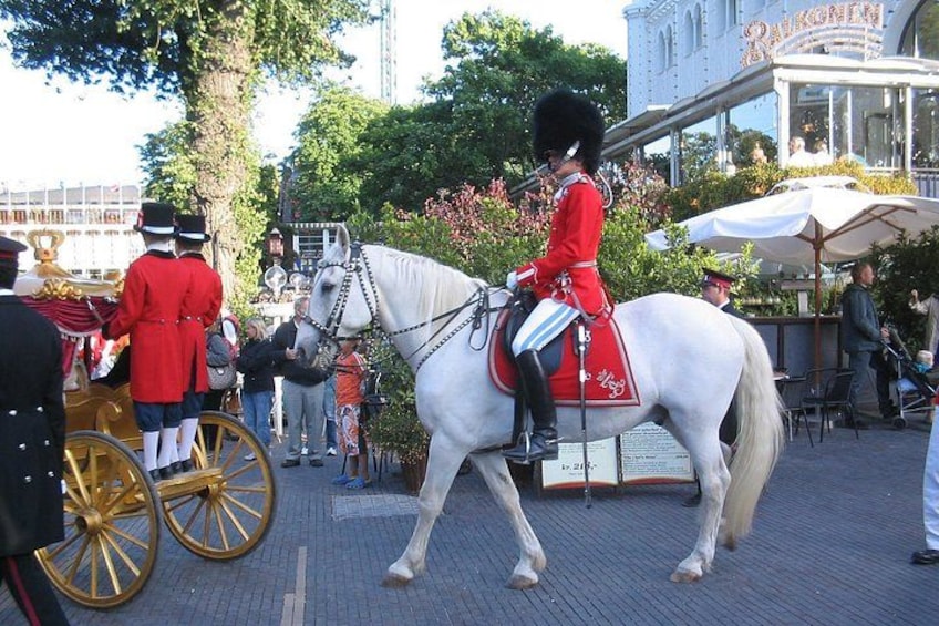 Una persona con uniforme rojo montando un caballo blanco con silla de montar roja, caminando sobre una superficie pavimentada con gente y un edificio al fondo. 3