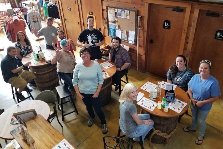 A group of people in a rustic interior with wooden tables and barrels, some seated and some standing. 3