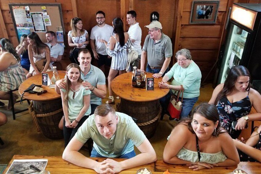 A group of people seated at wooden tables in a rustic indoor setting with barrels and a refrigerator. 4