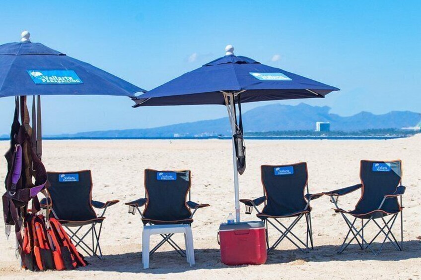 Beach chairs, umbrellas, and a cooler on a sandy beach with mountains in the background. 3
