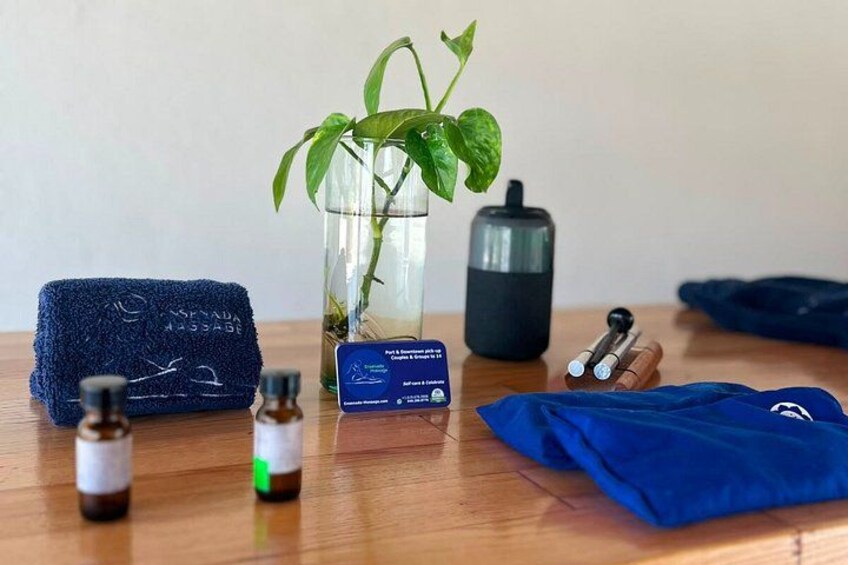 A wooden table with a blue towel, a glass vase with a plant, a black container, two small bottles, and a blue card. 4