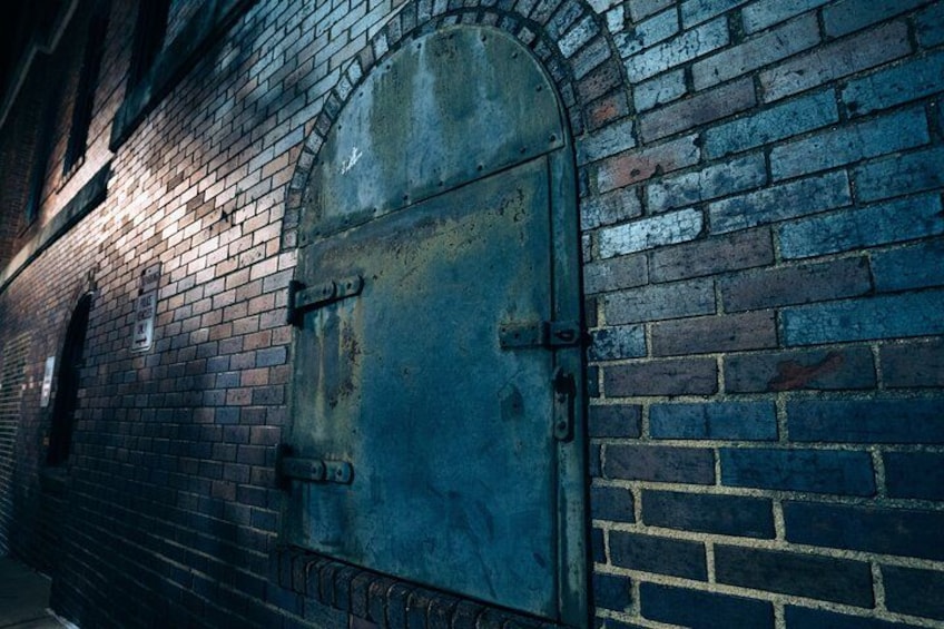 A large metal door with a metal latch, set in a brick wall with a window above it. 4