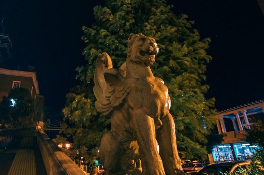 Una estatua de un león con una figura humana al fondo, un árbol y un edificio. 5
