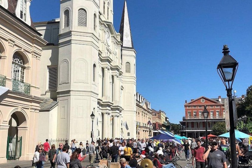 A crowded public square with historic buildings, including a church with a tall spire. 1