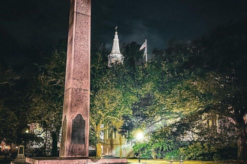 A night scene with a tall monument, a church spire, and surrounding trees. 4