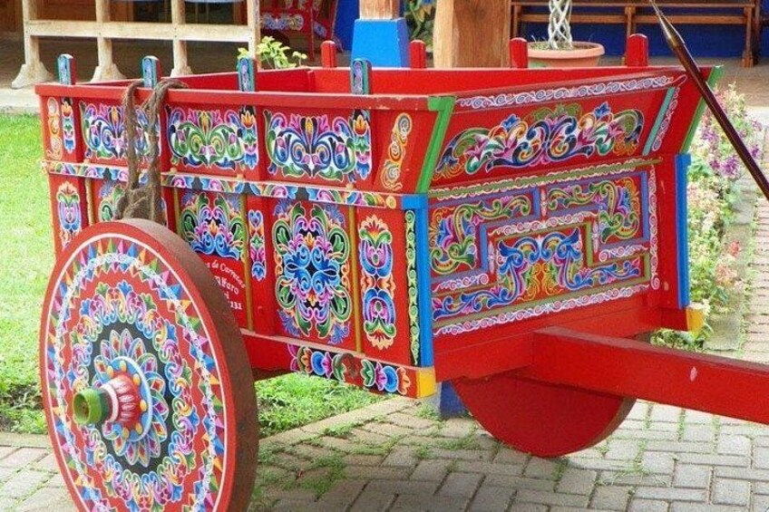 Un carretto colorato e finemente decorato, dotato di una grande ruota, posato su una superficie pavimentata con vegetazione sullo sfondo. 3
