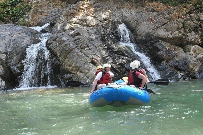 Een groep mensen die op een blauwe opblaasbare raft stroomafwaarts gaat in een rivier, met een waterval op de achtergrond. 3