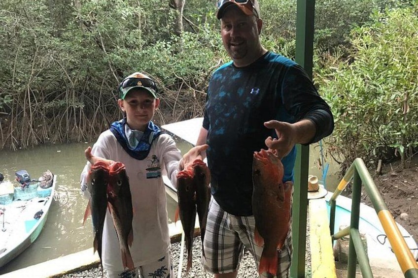 Two people holding fish, a boat, and a dock in a wooded area. 3