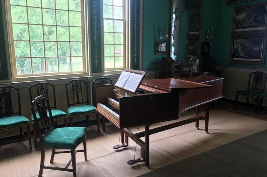 A grand piano in a room with large windows, green chairs, and framed pictures on the walls. 3