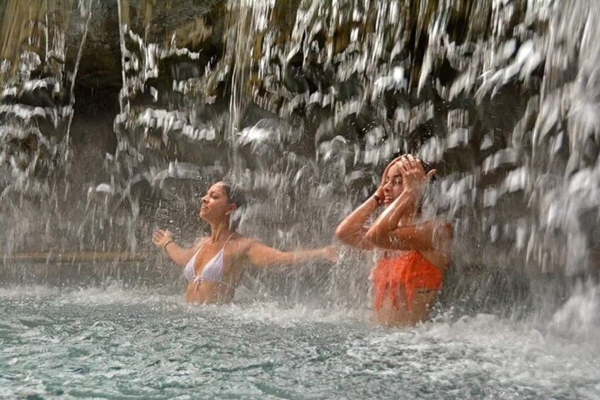 Two people in a water body with water cascading down a rocky background. 2