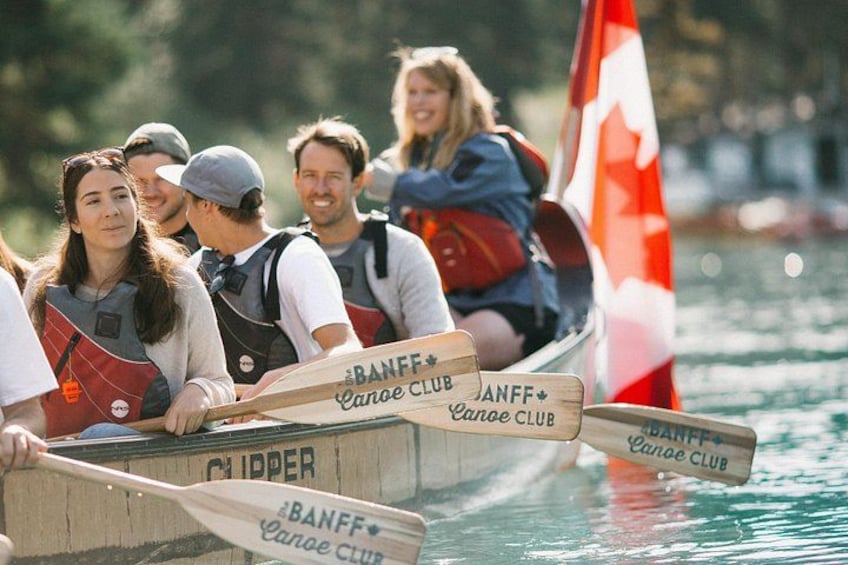 A group of people in a canoe on water, wearing life jackets and paddles with 'BANFF Canoe Club' written on them. 5