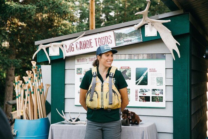 A person standing at a booth with a sign that reads 'BIG CANOE TOURS' and a wildlife spotting board. 3