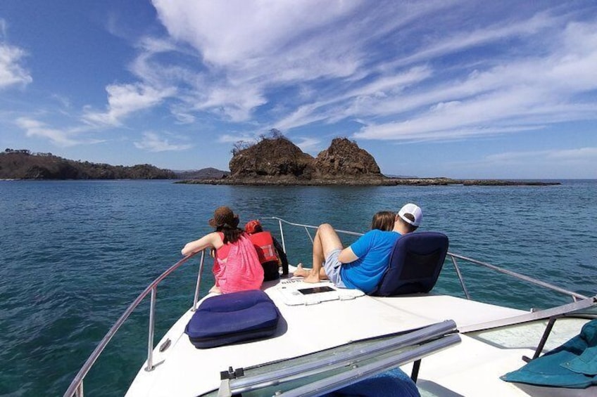 A boat with two people on deck, a rocky island in the distance, and calm water. 1