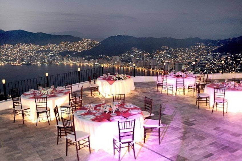 Un espacio de comedor al aire libre con mesas redondas dispuestas para una comida, con vista a un paisaje urbano al atardecer. 4