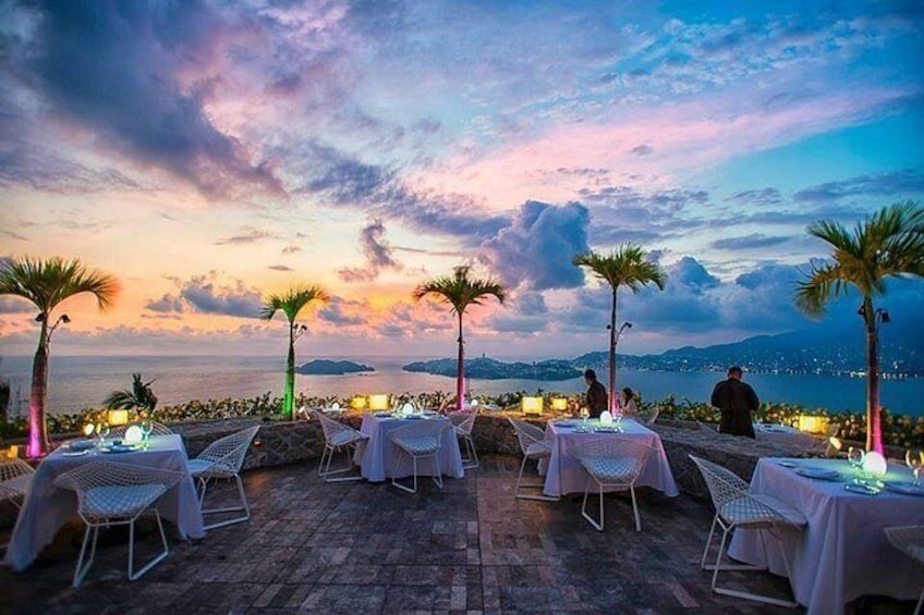 Un área de comedor al aire libre con mesas y sillas, con vista a un hermoso atardecer sobre el mar. 1