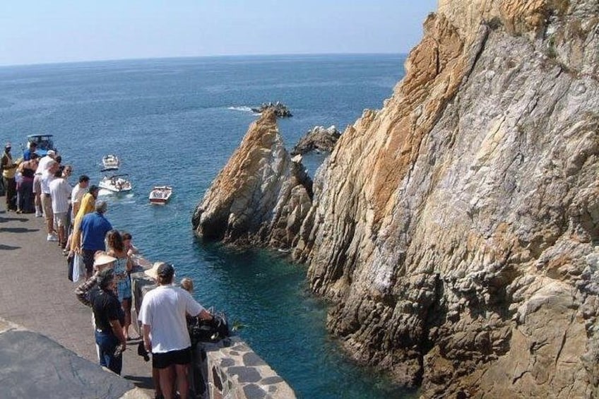 A rocky coastline with people standing on a path overlooking the sea, boats in the water, and a clear sky. 1