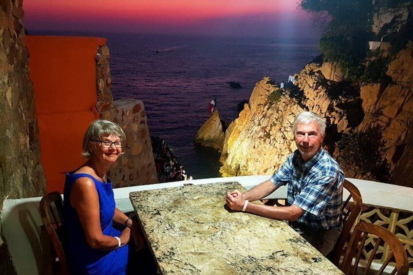 A couple seated at a table overlooking a rocky coastline with the ocean in the background. 1
