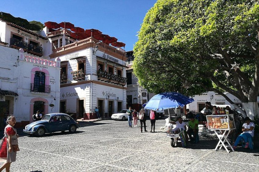 A cobblestone street with buildings, a tree, and people walking. 1