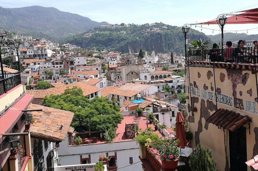 A densely packed urban area with terracotta-roofed buildings, a prominent building with a sign, and a mountainous backdrop. 3
