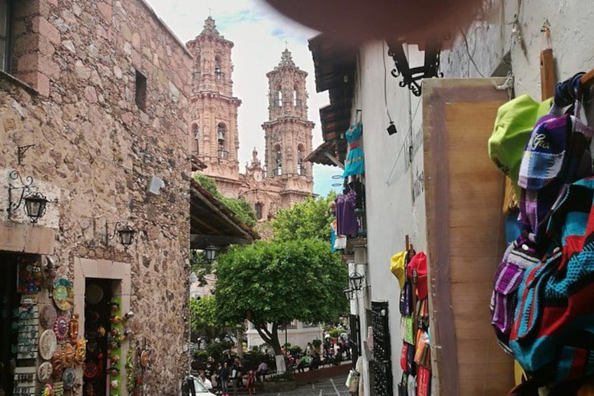 A narrow street with colorful textiles hanging on a wall, a stone building, and a bell tower in the background. 5