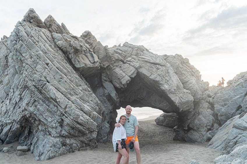 Una pareja parada frente a una gran formación rocosa en forma de arco sobre una playa arenosa. 4