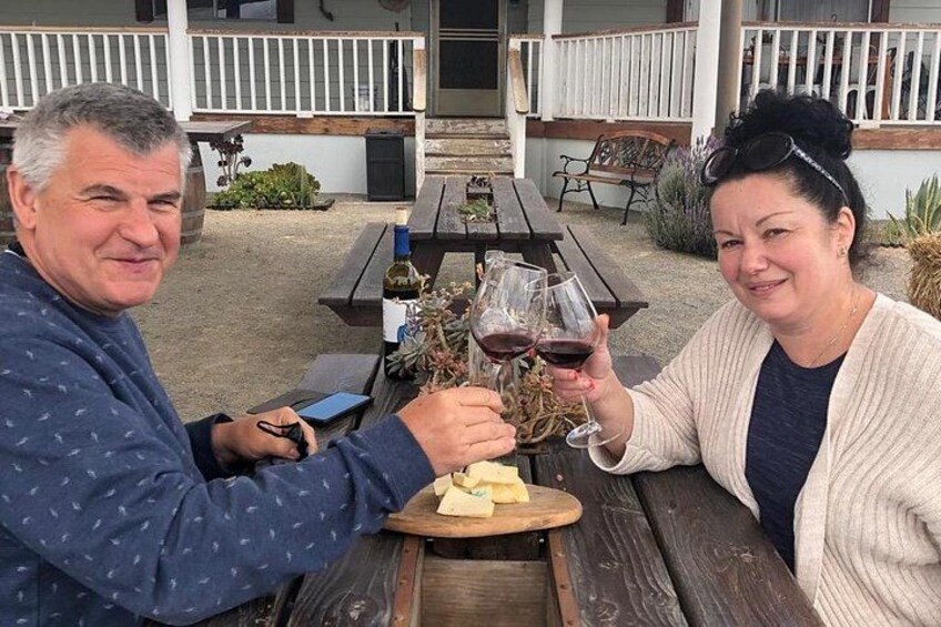 Two people toasting with wine glasses at an outdoor table with a wooden bench, surrounded by a white fence and a building with a porch. 4