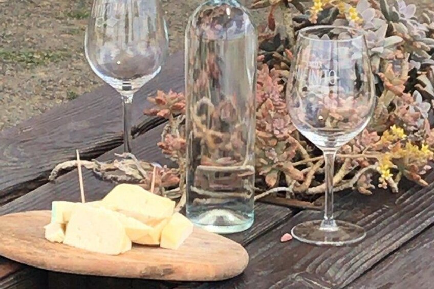 A wooden cutting board with cheese, a glass bottle, and two wine glasses on a wooden table with dried flowers. 2