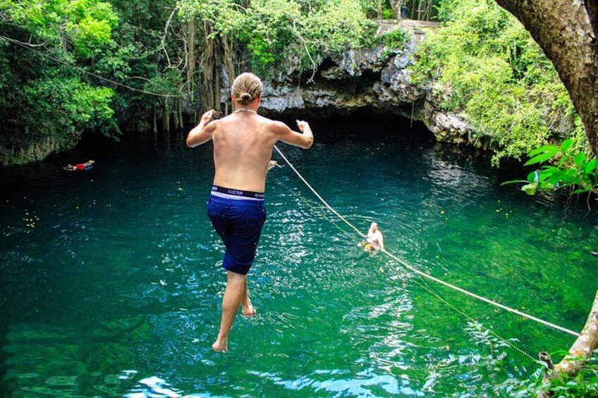 Une personne se tient debout sur une corde tendue au-dessus de l’eau, une autre personne se trouve dans l’eau, et une grotte est visible au fond. 2