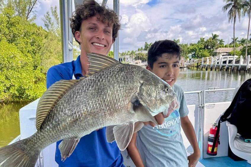 Two people holding a large fish on a boat with a waterway and palm trees in the background. 1