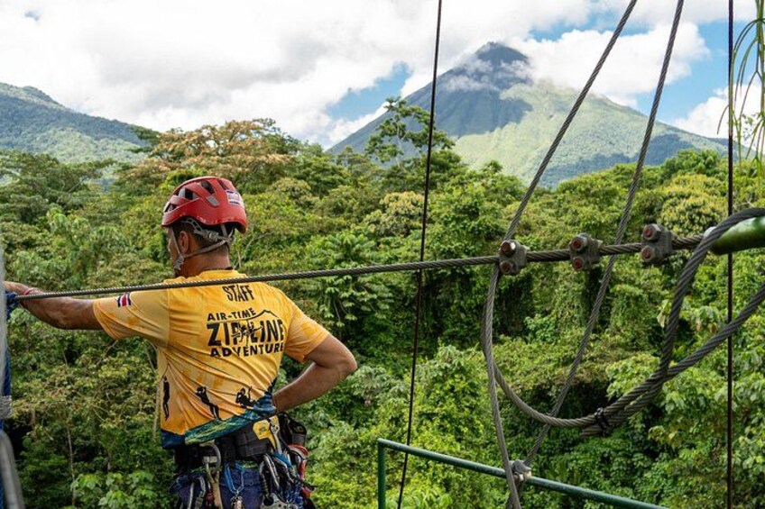 Een medewerker van een avonturenpark, met een geel shirt en rode helm, kijkt uit over een weelderig groen bos en een berg op de achtergrond. 1