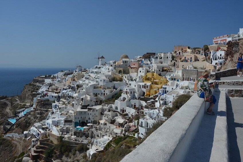 A coastal village with white and yellow buildings, a windmill, and a person sitting on a ledge. 2