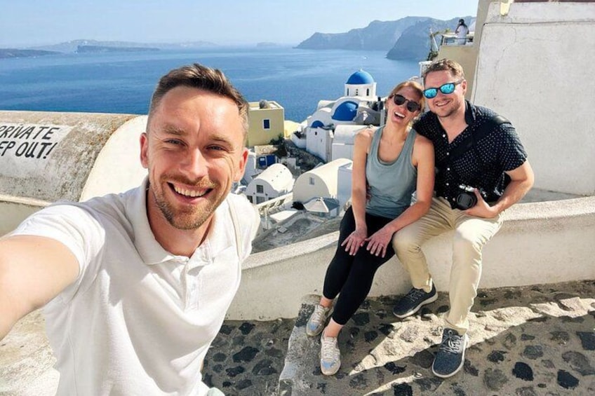A person taking a selfie with a scenic view of a coastal area, including white buildings and the sea. 2