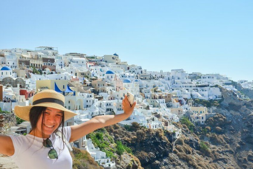 A person with a straw hat and sunglasses, standing on a cliff overlooking a white-washed village with blue domes, set against a clear sky. 4