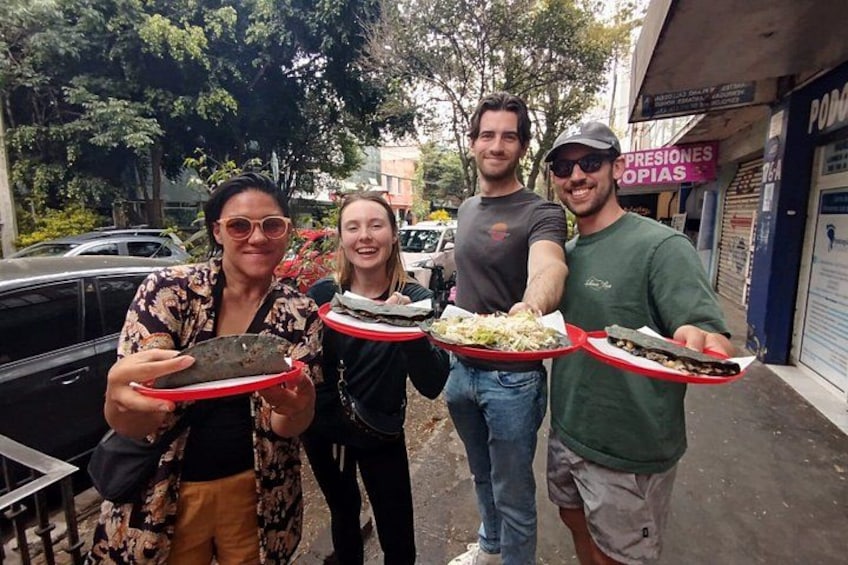 Four people holding food items on red trays, standing on a street with cars and a building in the background. 1