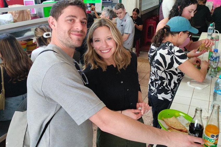 A man and a woman standing in a busy indoor setting, possibly a cafe or restaurant, with other people in the background. 4