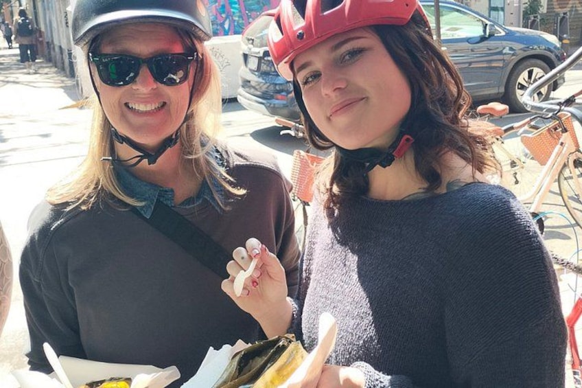 Two people wearing helmets and holding food items, with bicycles and a car in the background. 2