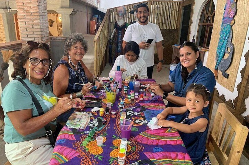 A group of people sitting around a table with a colorful tablecloth, engaged in painting activities. 1