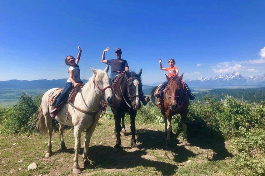 Three people riding horses on a grassy hill with a mountainous landscape in the background. 3