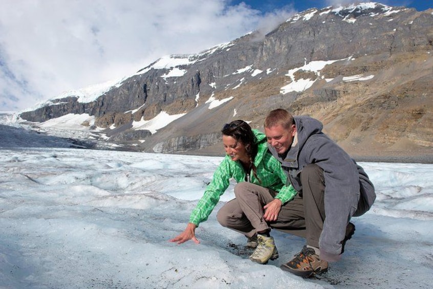 Two people crouching on a glacier with mountains in the background. 2